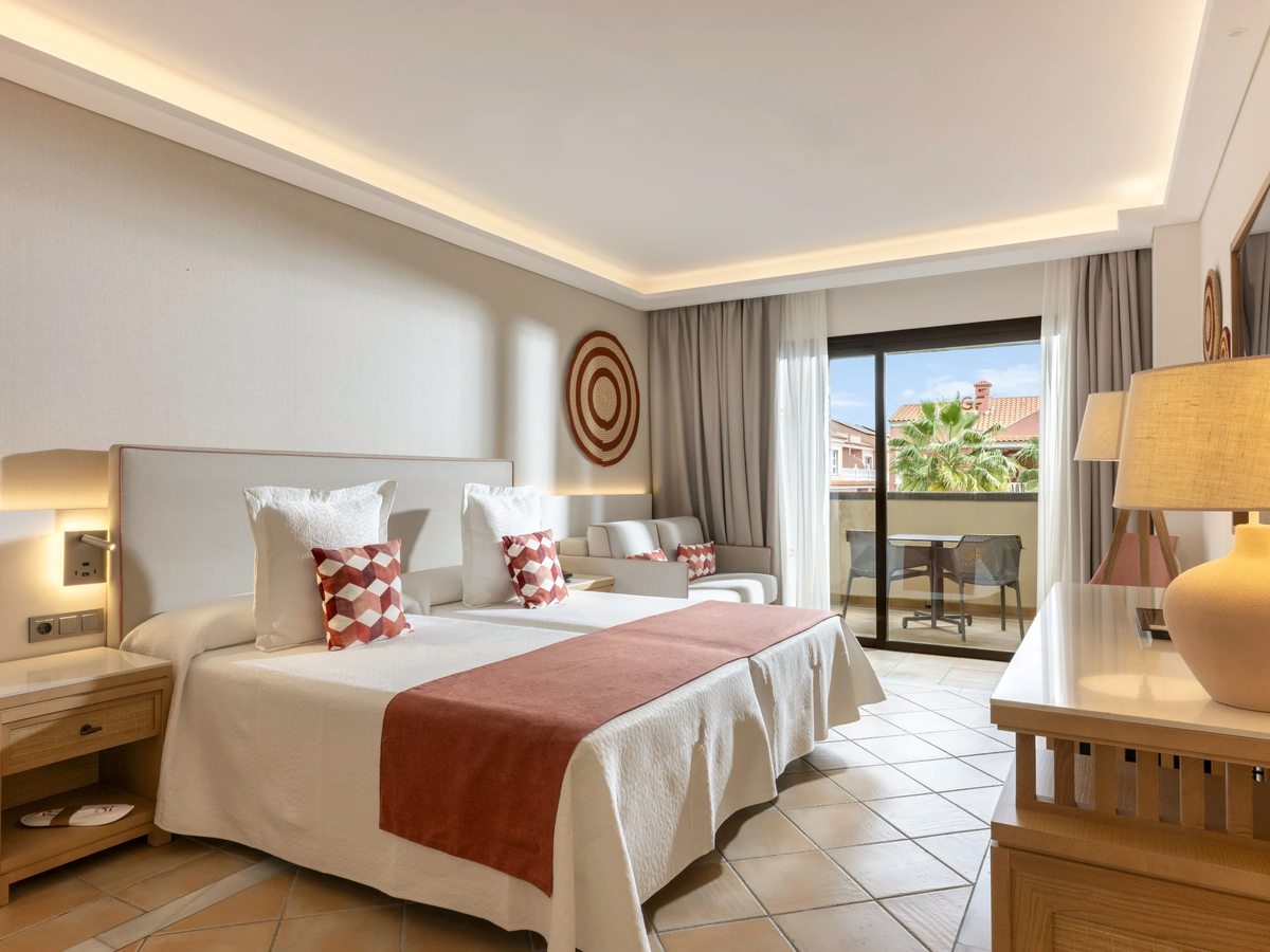 A bright room with two single beds pushed together under a rust-red runner, a sofa, a desk and a balcony beyond.