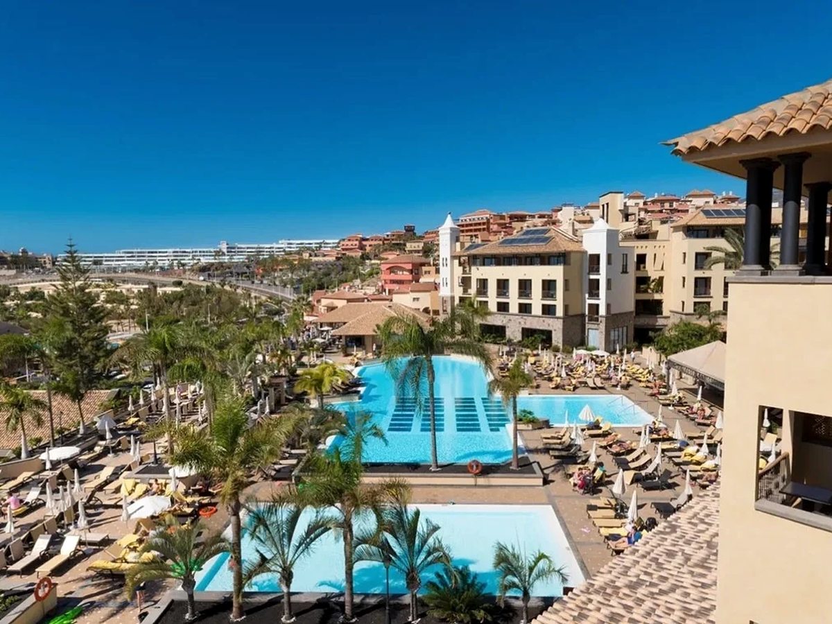 An elevated view over the resort's two main pools, rows of loungers and parasols, palm-planted terrace and the hotel buildings behind.