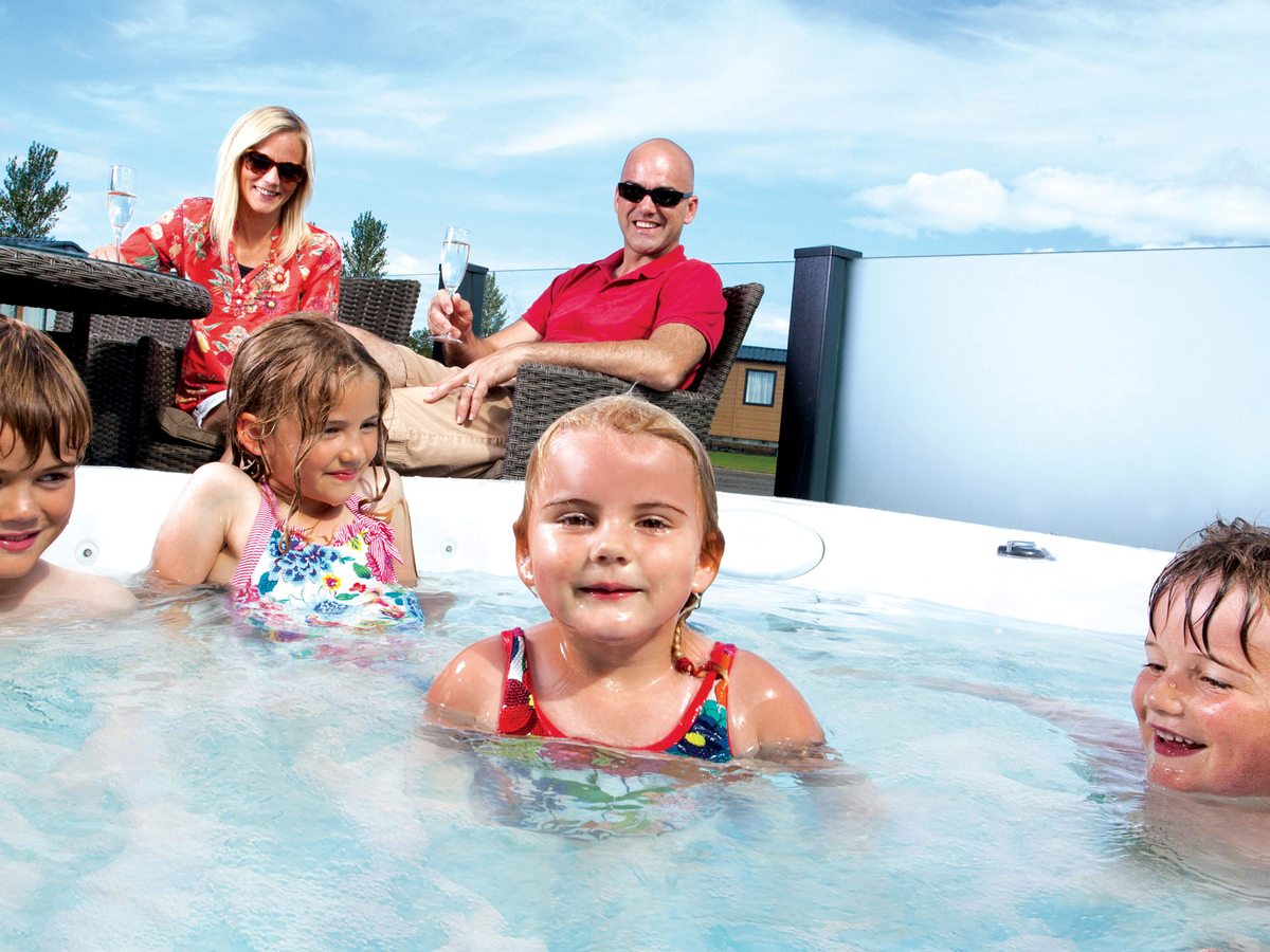 Children playing in a bubbling hot tub while two adults sit behind with drinks, lodges and blue sky beyond