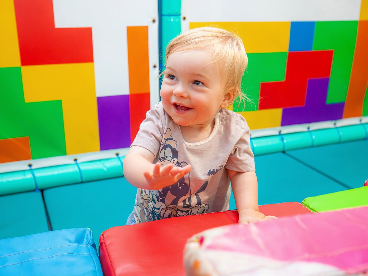 A blond toddler smiling among the colourful padded blocks of the indoor soft play