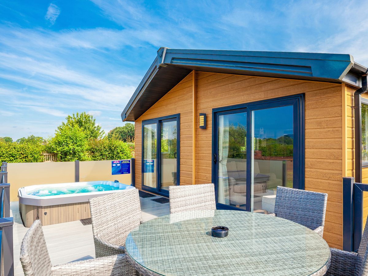A timber-clad lodge with sliding doors and a decked terrace holding a bubbling hot tub and a table and chairs, under a blue sky