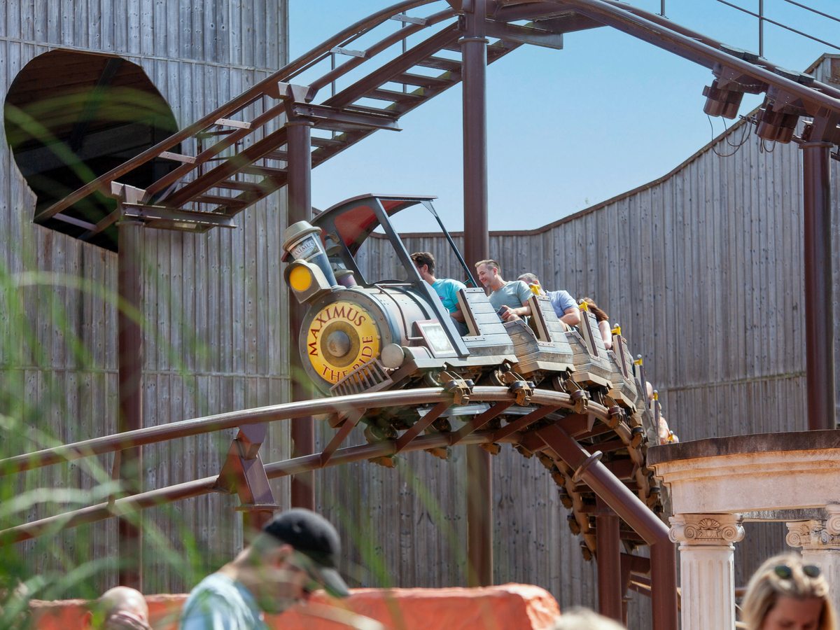 The Maximus locomotive-fronted rollercoaster train rounding a bend with riders aboard, under a blue sky
