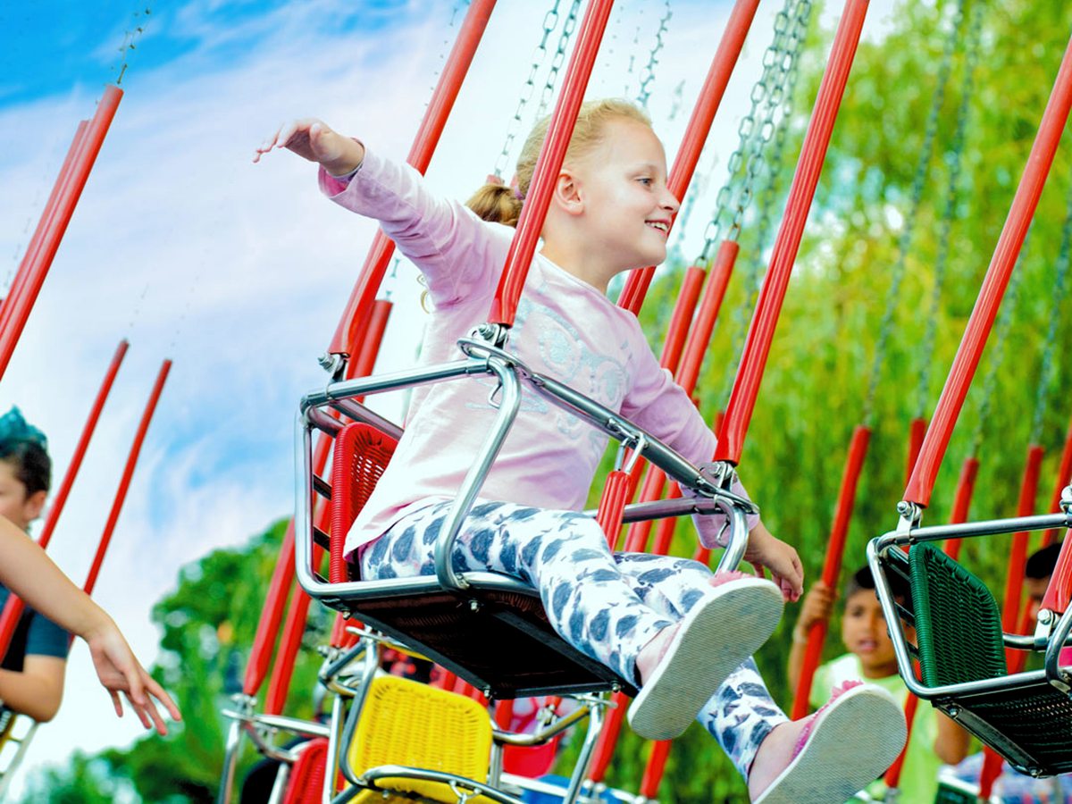 A young girl smiling on a chain swing seat of the swing ride, one arm outstretched, green trees behind