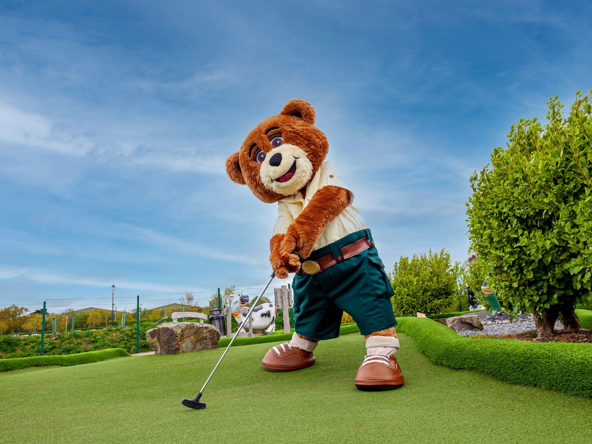 The Crealy bear mascot lining up a putt on a landscaped mini-golf green under a blue sky