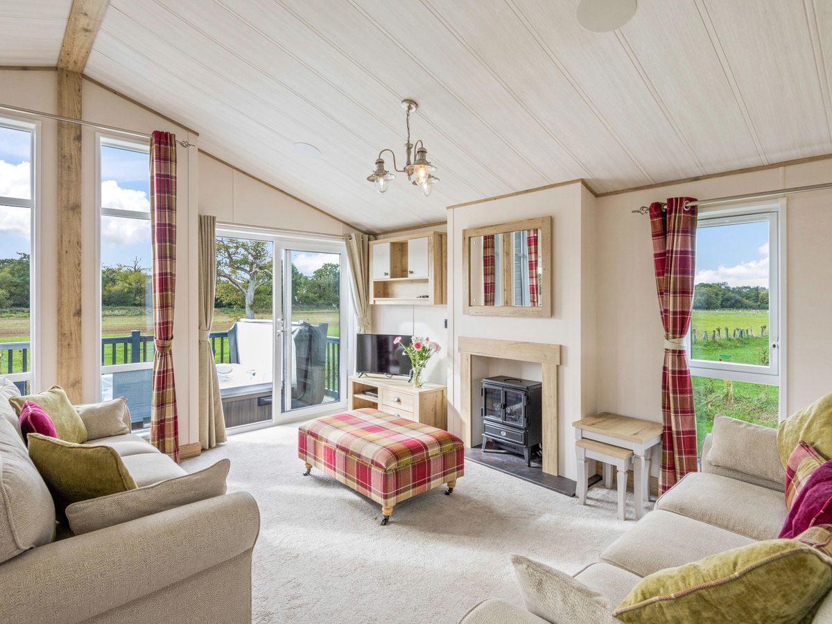 A lodge lounge with cream sofas, a tartan footstool, a stove-style fireplace and patio doors to the deck