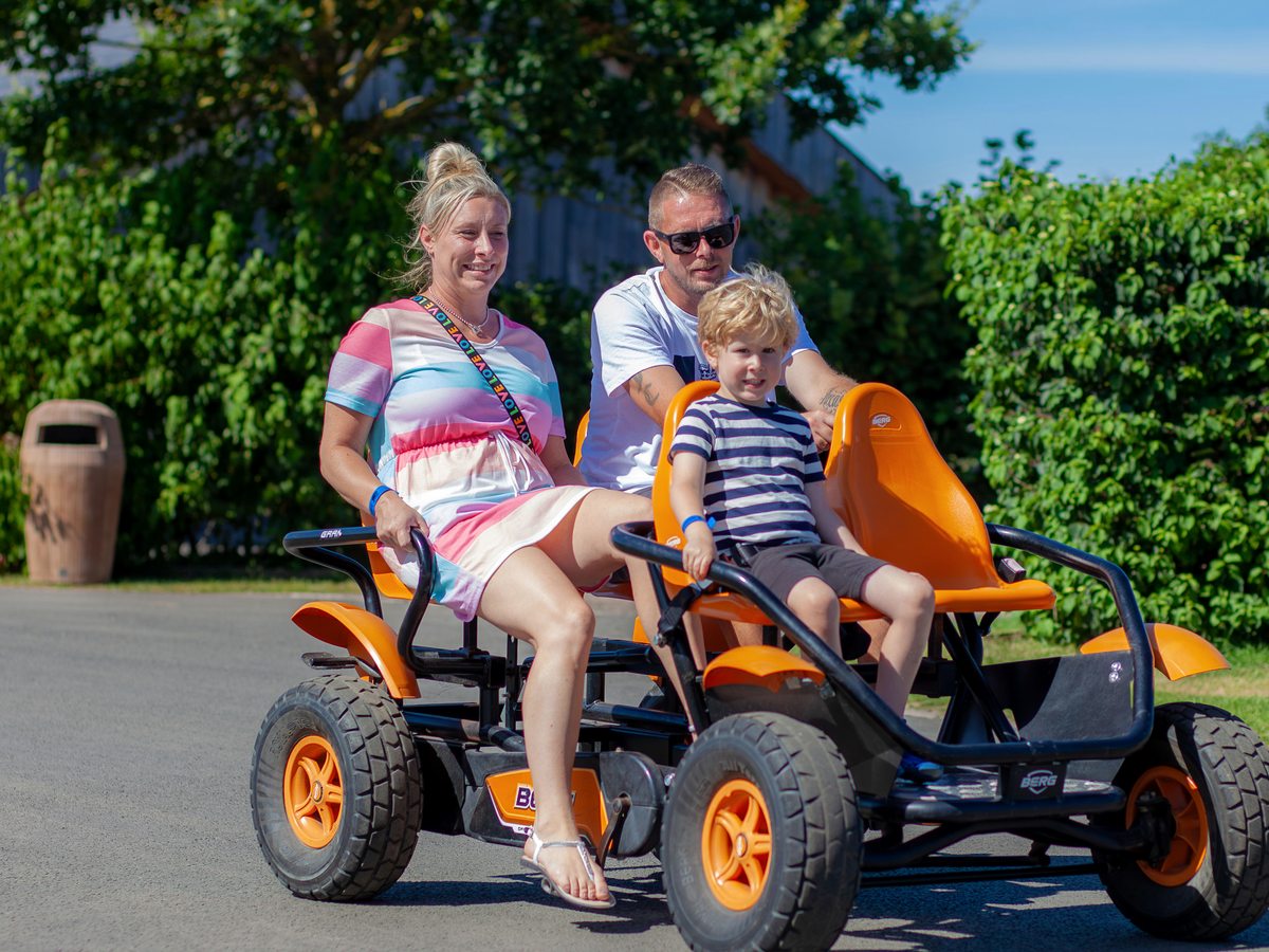 A family riding an orange four-wheeled Berg pedal kart along a tarmac path lined with hedges
