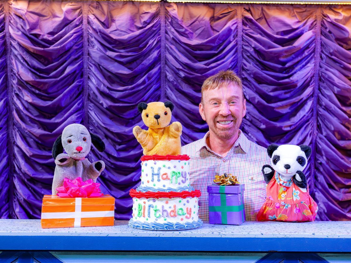 A performer on a purple-curtained stage with Sooty, Sweep and Soo puppets around a Happy Birthday cake