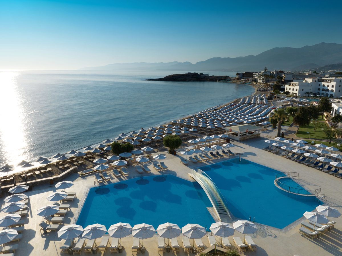 Aerial view of the resort's two large pools in the foreground, rows of loungers along a sandy beach and the bay and mountains beyond