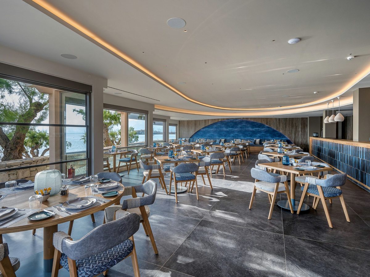 A modern restaurant interior with wooden chairs and grey upholstery, tables set beside floor-to-ceiling windows onto the sea and a curved blue feature wall