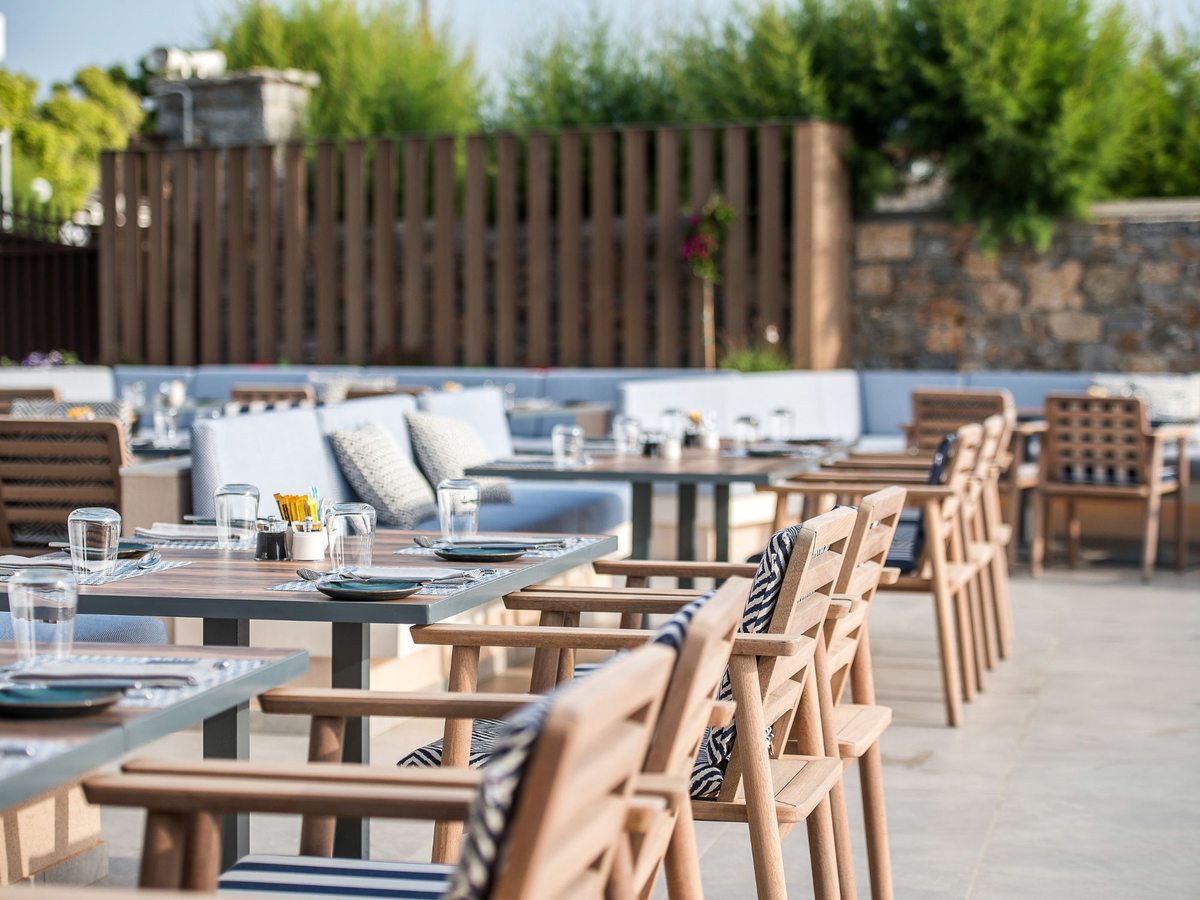 An outdoor restaurant terrace with wooden armchairs, pale banquettes and set tables beside a timber screen and stone wall