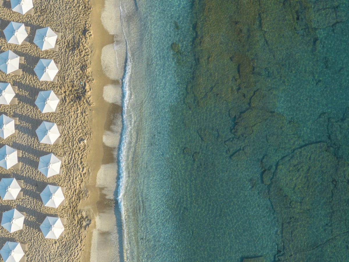 Aerial view of the resort beach, rows of white umbrellas on pale sand meeting clear shallow water over sand and rock