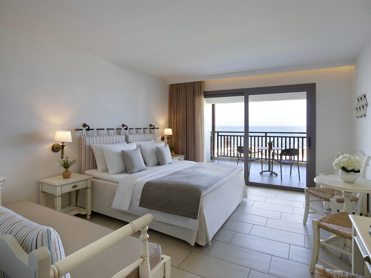 A bright bedroom with a double bed, a striped headboard and a small sofa, with sliding doors onto a balcony with a sea view