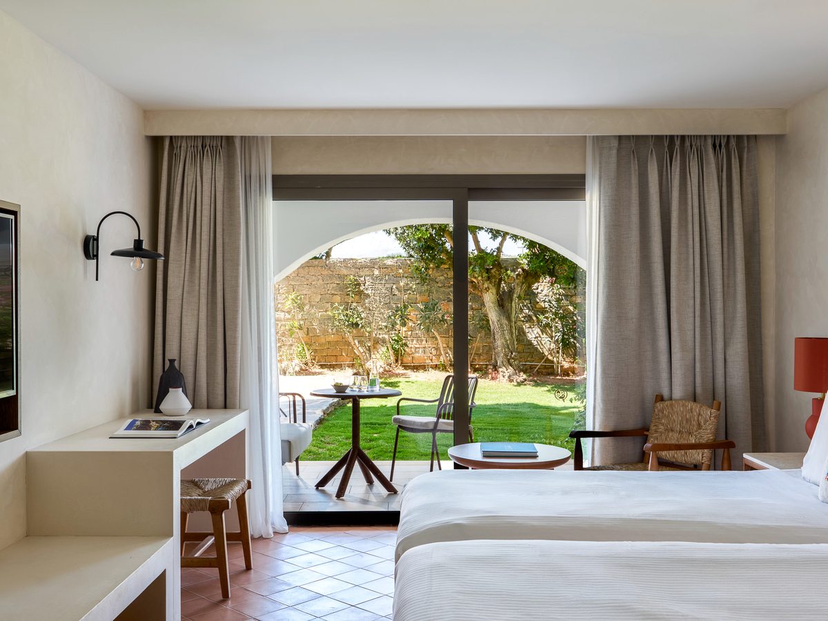 A room with twin beds and a desk, sliding doors opening onto a private lawn behind an arched stone garden wall