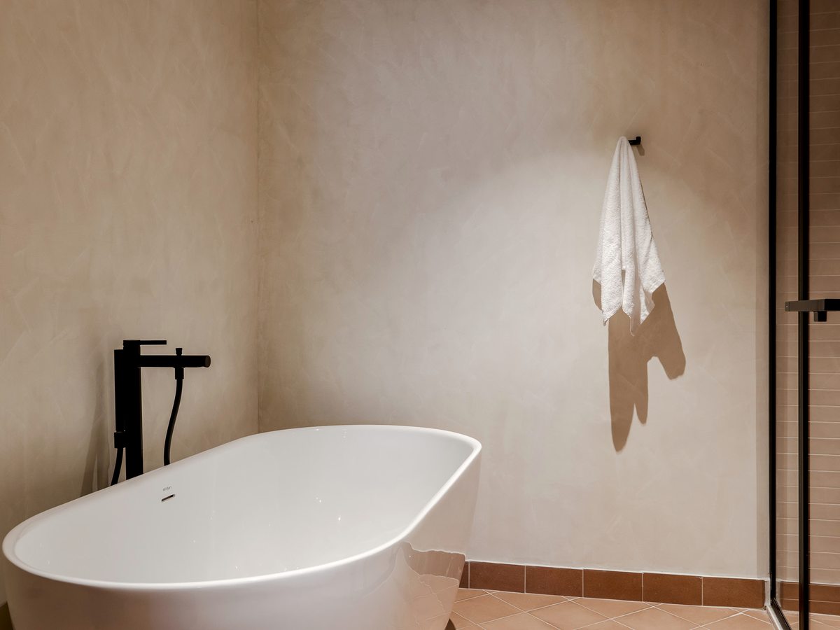 A bathroom finished in pale microcement with a white freestanding bath and a glass shower screen