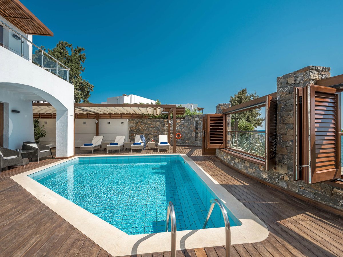 A private rectangular pool on a wooden deck with sun loungers and a pergola, with a sea view beyond a stone wall
