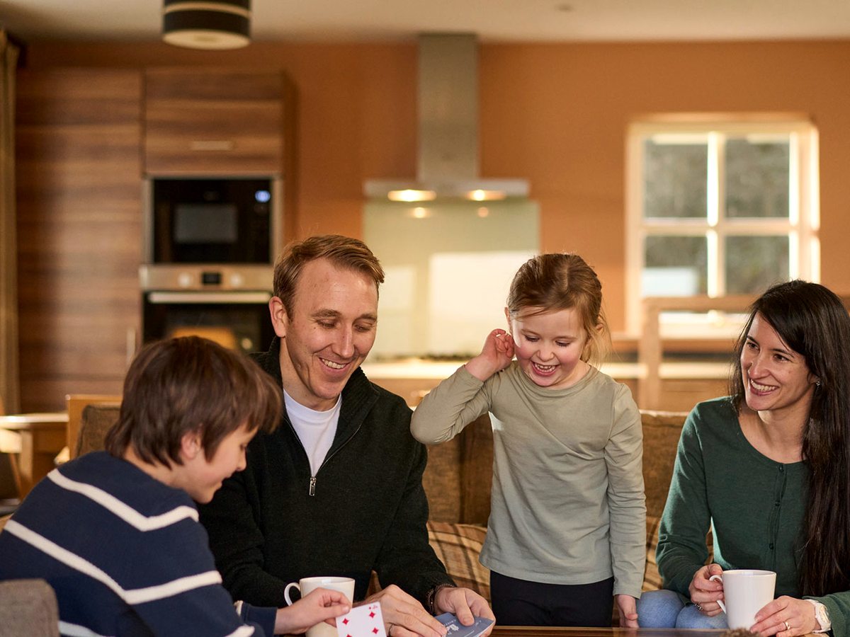 A family sitting and playing cards on a sofa in a lodge living room, with an open-plan fitted kitchen and oven behind.