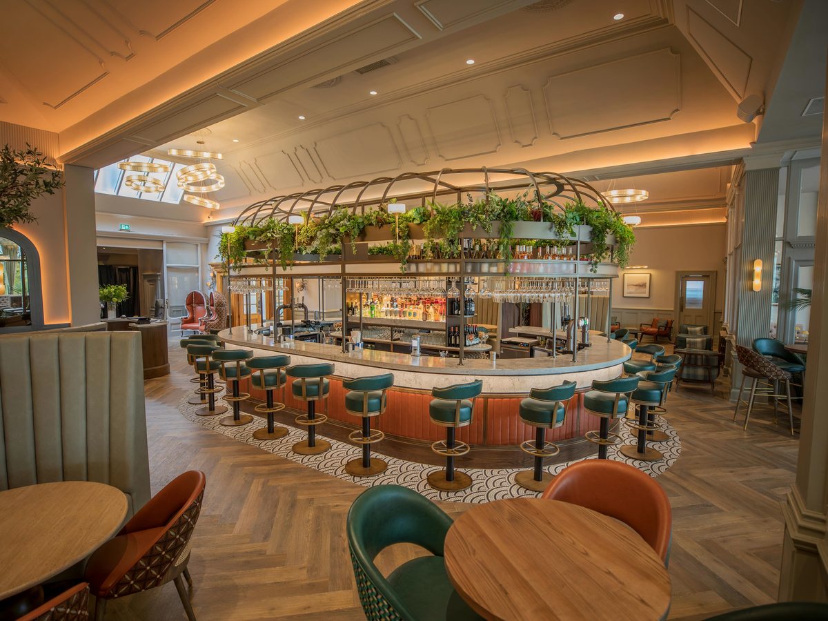 An oval island bar with green leather stools and a coral-panelled base, hanging glassware and trailing plants above, in a panelled room with herringbone flooring.