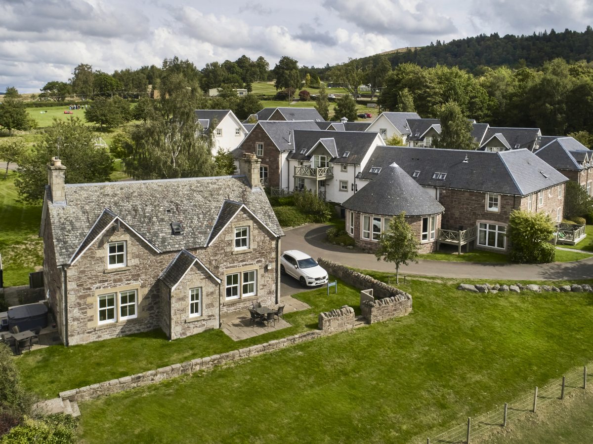 An aerial view of stone-built holiday lodges and cottages with slate roofs set among lawns and trees, with wooded hills behind.