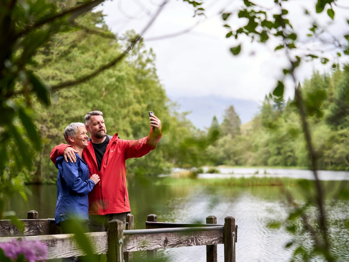 Two people on a wooden-railed lochside path in the estate grounds, with still water, trees and hills around them.