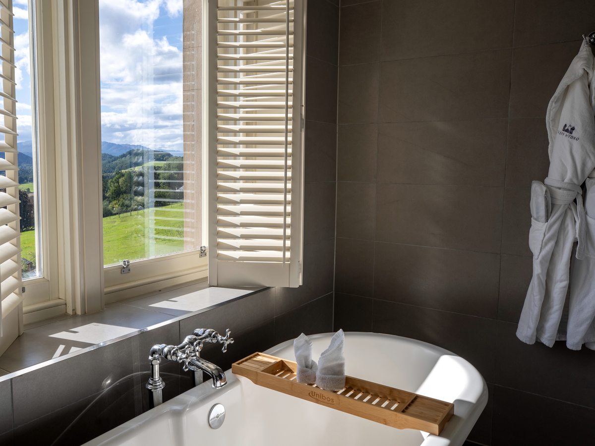 A freestanding bath beside a shuttered window looking onto a green lawn, with dark wall tiles and bathrobes alongside.