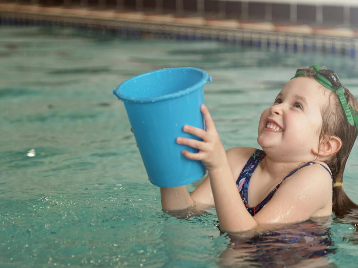 A young child in a swimsuit standing in the shallow pool water, smiling and holding up a blue plastic bucket.