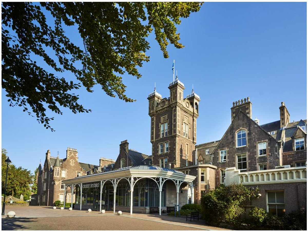 The red-sandstone Victorian hotel with its clock tower and a glass-roofed entrance canopy signed Crieff Hydro, under a clear blue sky.