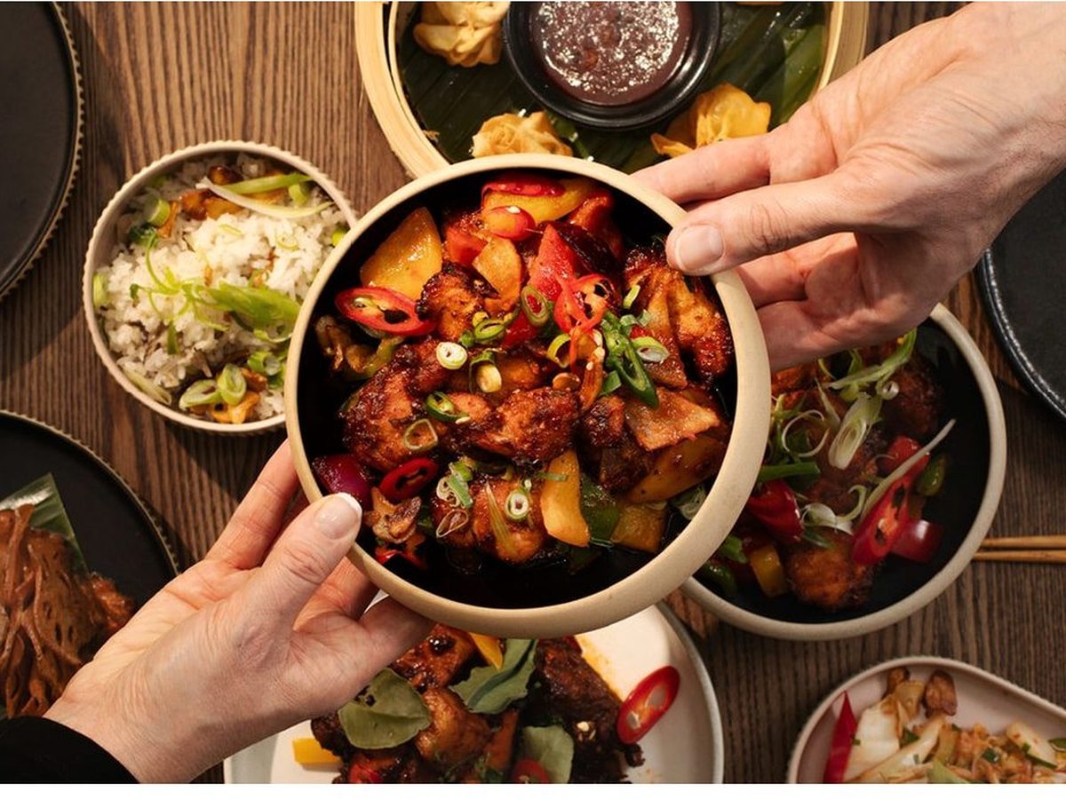 Hands lifting a bowl of a chunky Asian dish above a table set with rice, noodles and other small sharing plates.