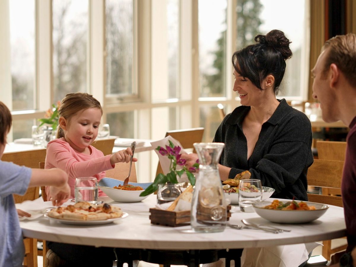A family of four at a round marble table by tall windows, with pizza and pasta bowls, the children reaching for food.