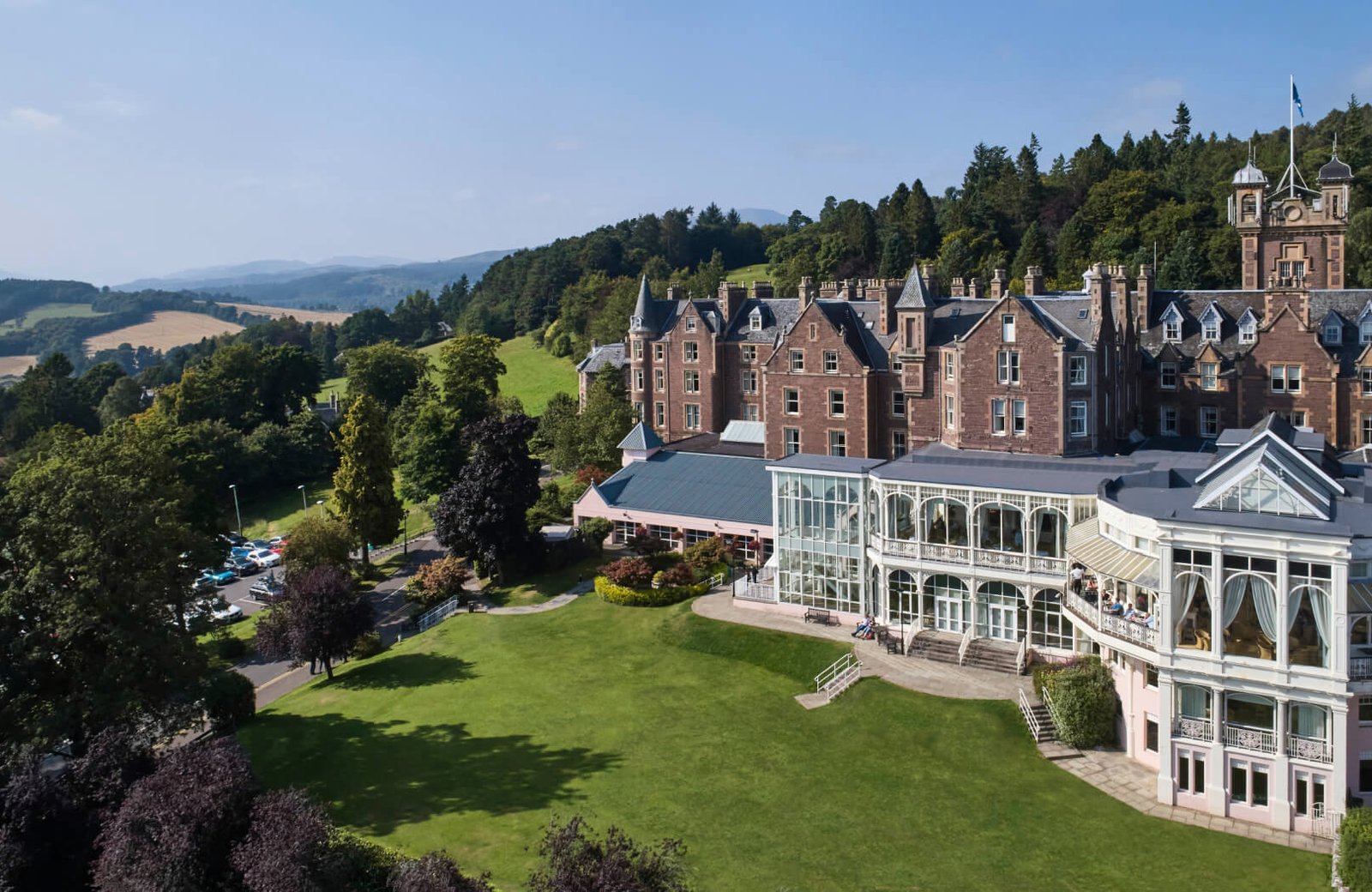 The red-sandstone Victorian hotel and its glass conservatory above a green lawn on the estate, with the Strathearn Valley and hills beyond.
