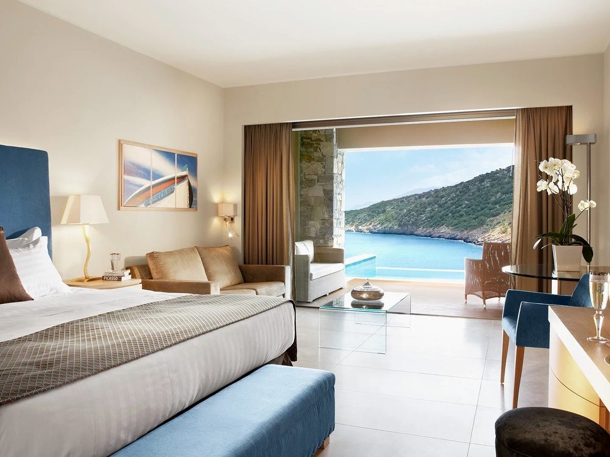 A double room with a bed against a wooden headboard, a bench seat and an open balcony door to a hillside and sea view.
