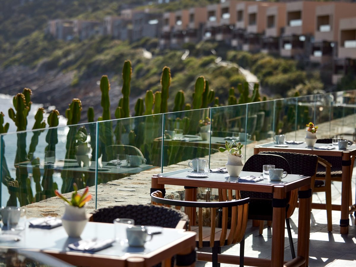 White-clothed tables set for breakfast on an open stone terrace, cactus planting to one side and the cove below.