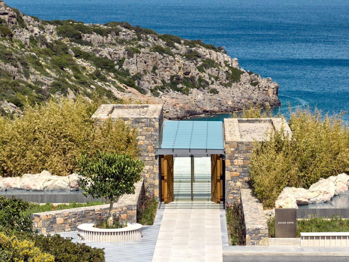 A glass and stone entrance building with a paved walkway and olive trees, the rocky coastline and sea beyond.