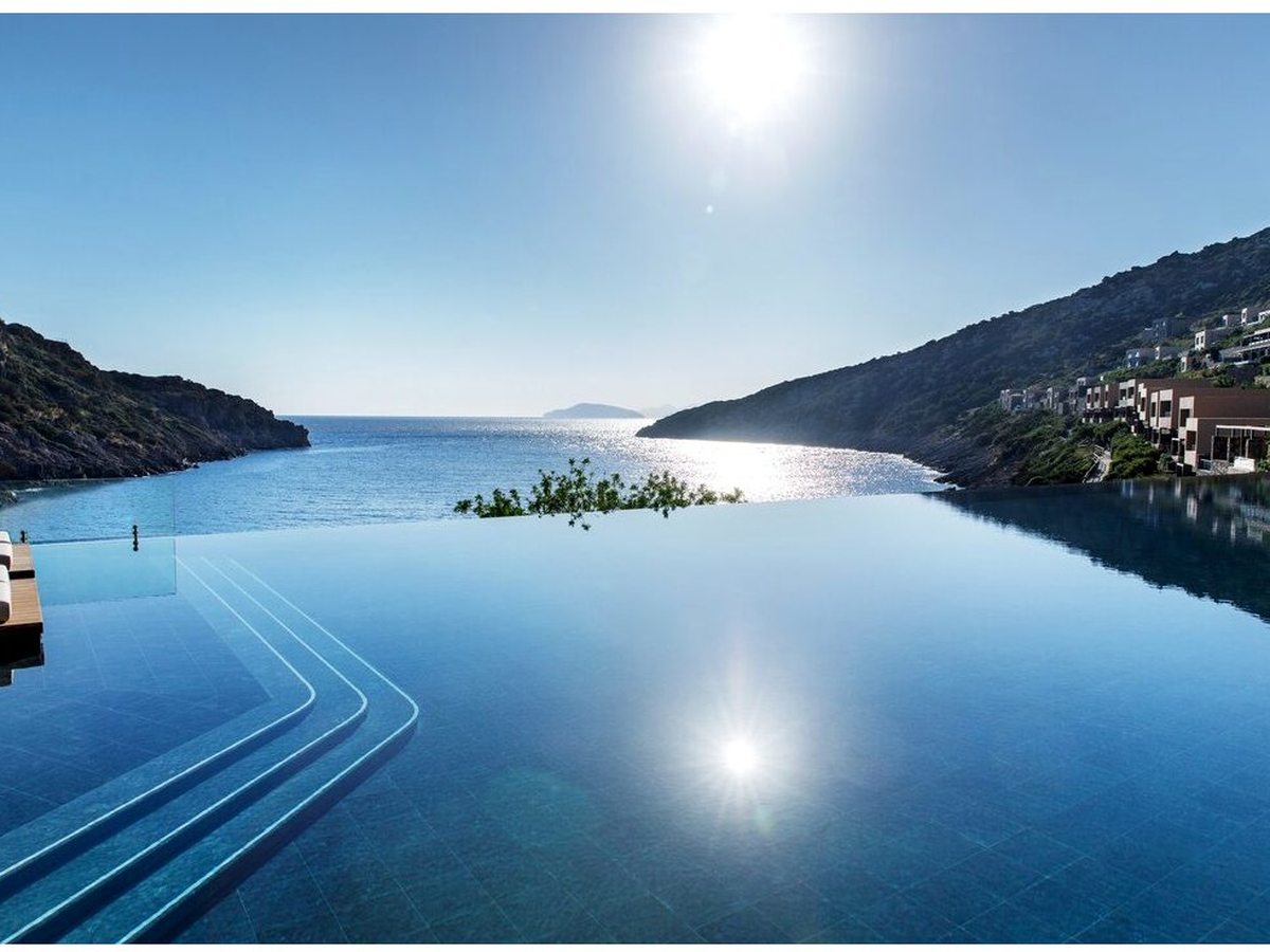A long infinity pool reflecting the midday sun, its far edge open to the sheltered bay and a rocky headland beyond.