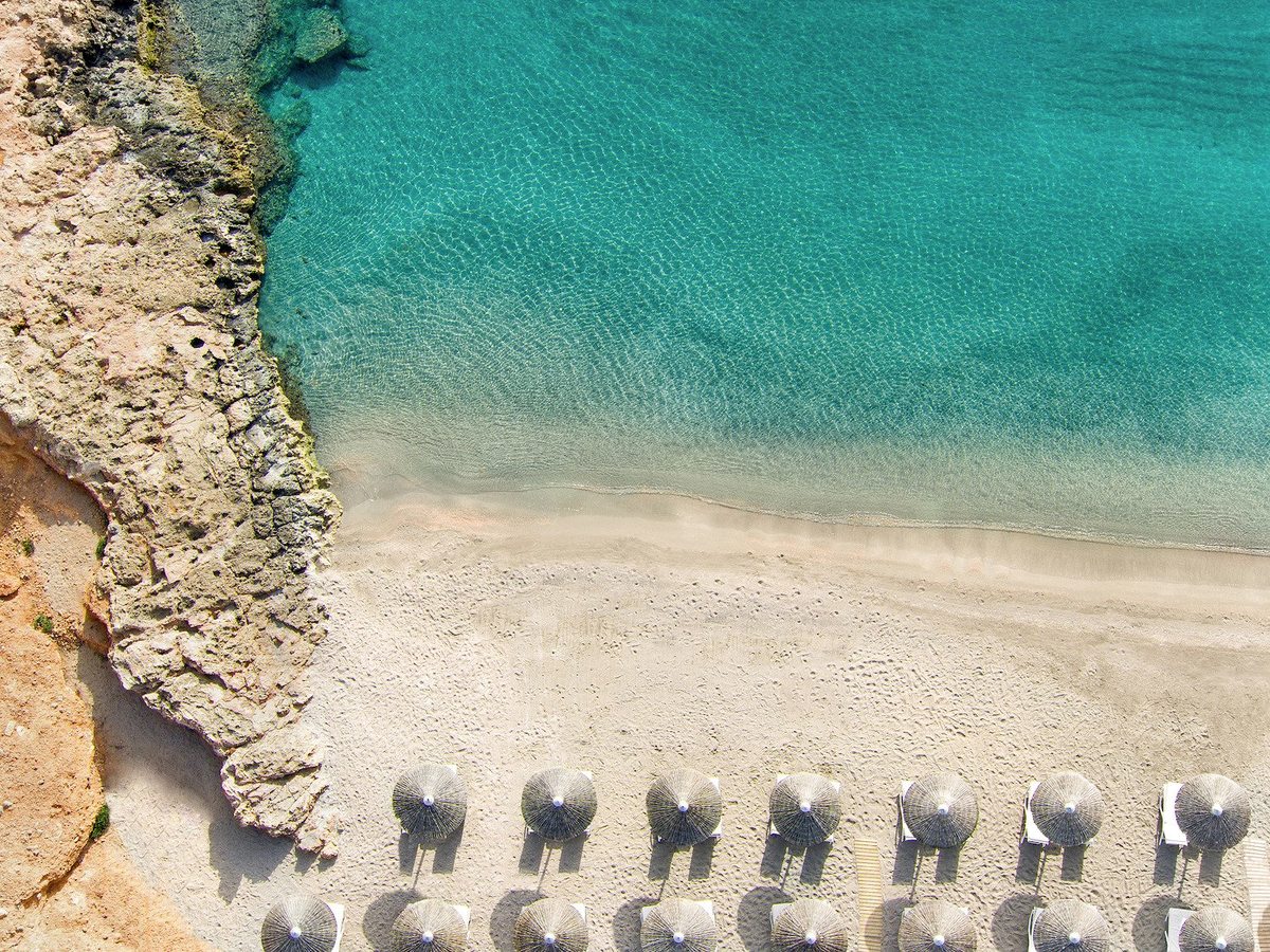 An overhead view of the sandy beach meeting clear turquoise shallows, with rows of grey parasols along the sand.