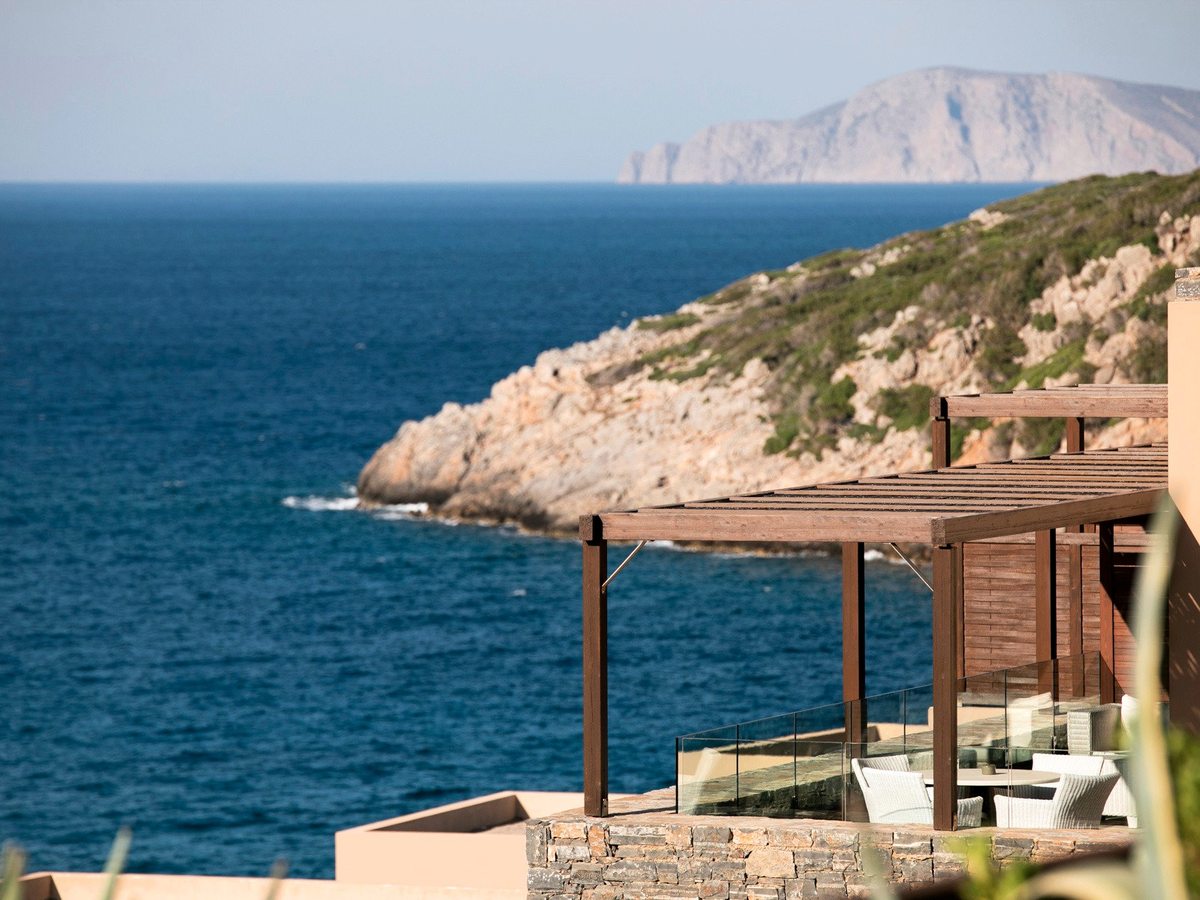 A terrace with a timber pergola and glass balustrade looking across the blue bay to a rocky headland and an offshore island.