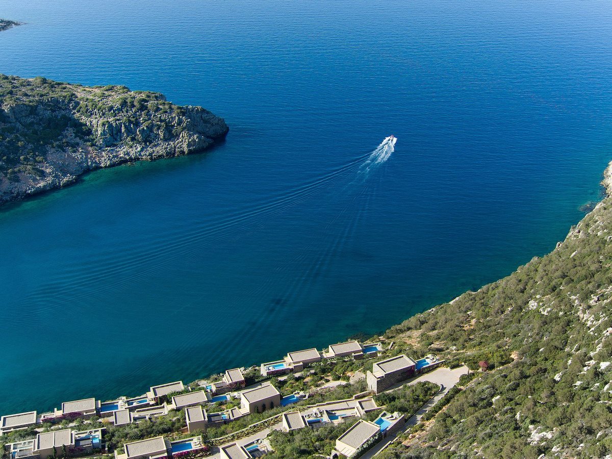An aerial view of the sheltered bay's rocky coastline and deep-blue water, a motorboat leaving a wake below the hillside resort.