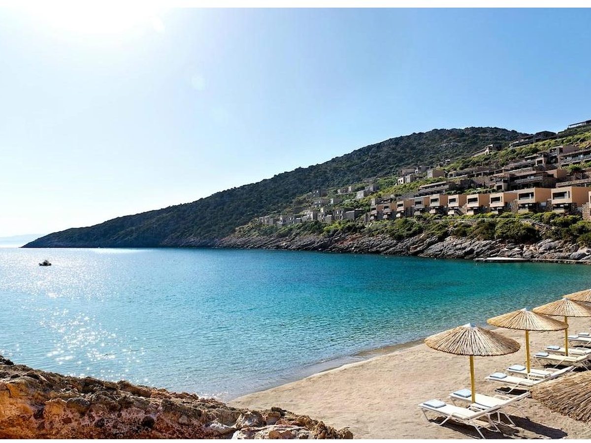 A sheltered sandy beach with straw parasols and sunbeds along the shore, clear shallow water and hillside villas behind.
