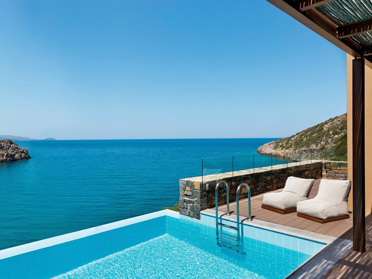 A private infinity pool with a timber deck and glass screen, its edge open to the turquoise cove and rocky coast.
