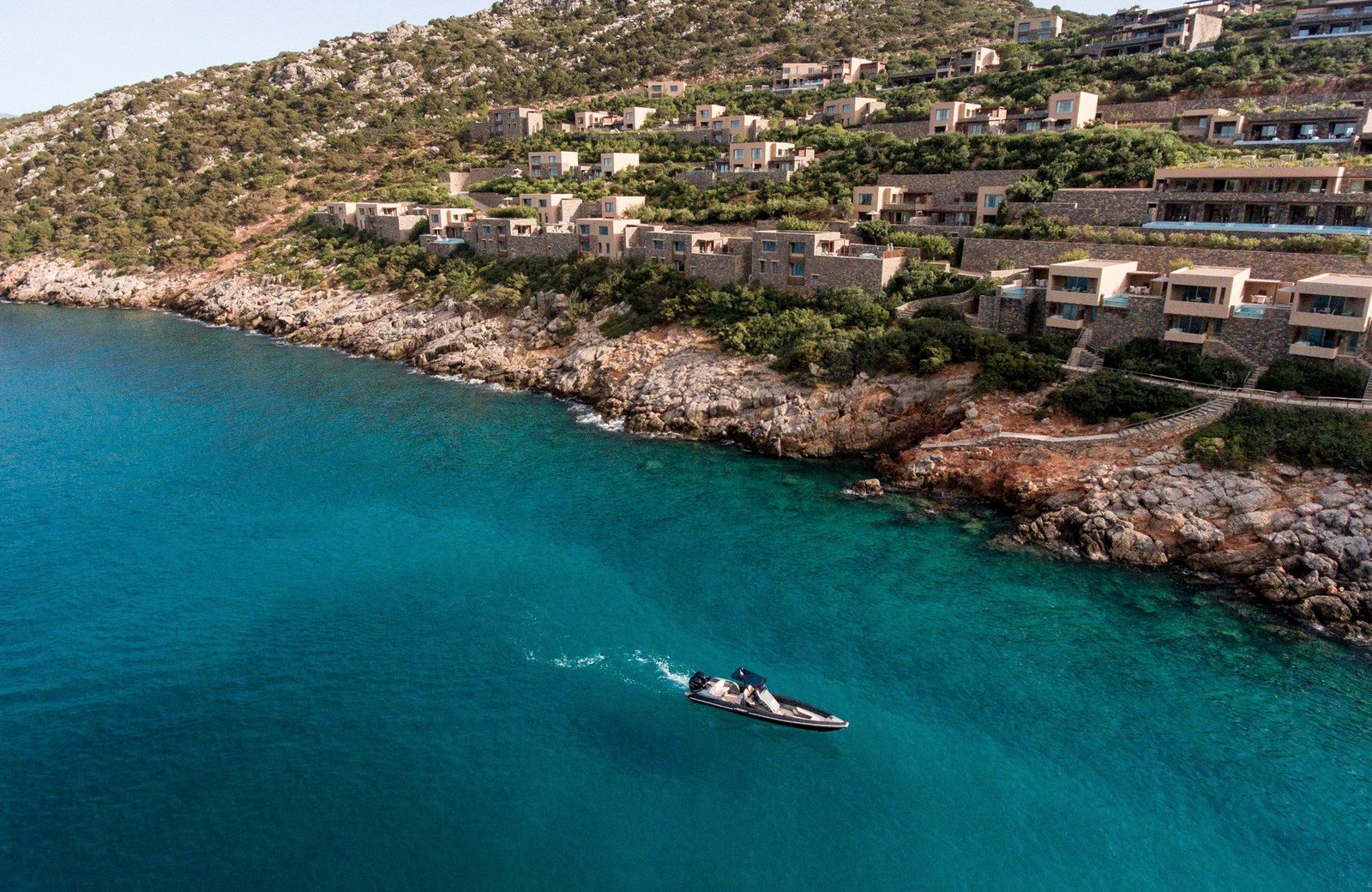 Stone villas stepping down a green hillside to a turquoise cove on Mirabello Bay in north-east Crete, a small boat on the water.