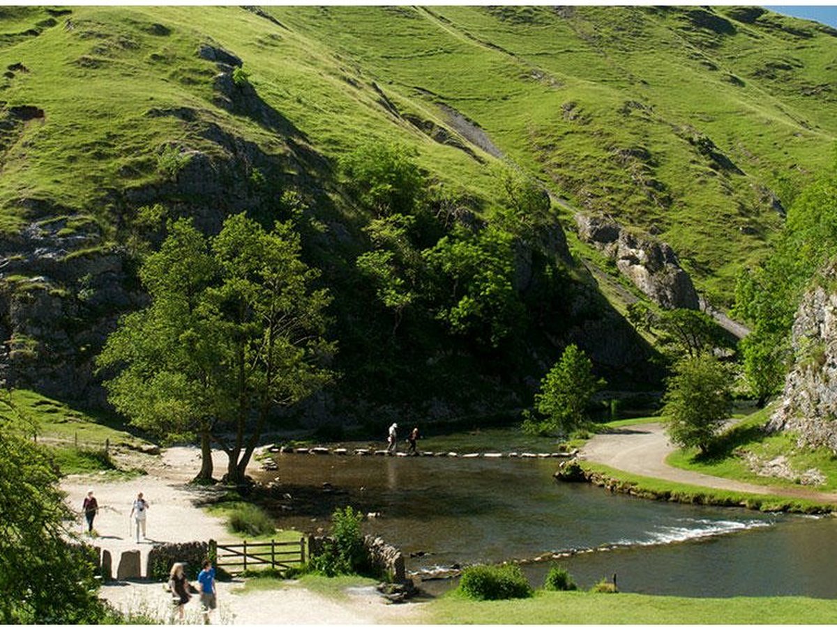 A green limestone valley in the Peak District with a shallow river, stepping stones and a footpath, walkers in the distance.