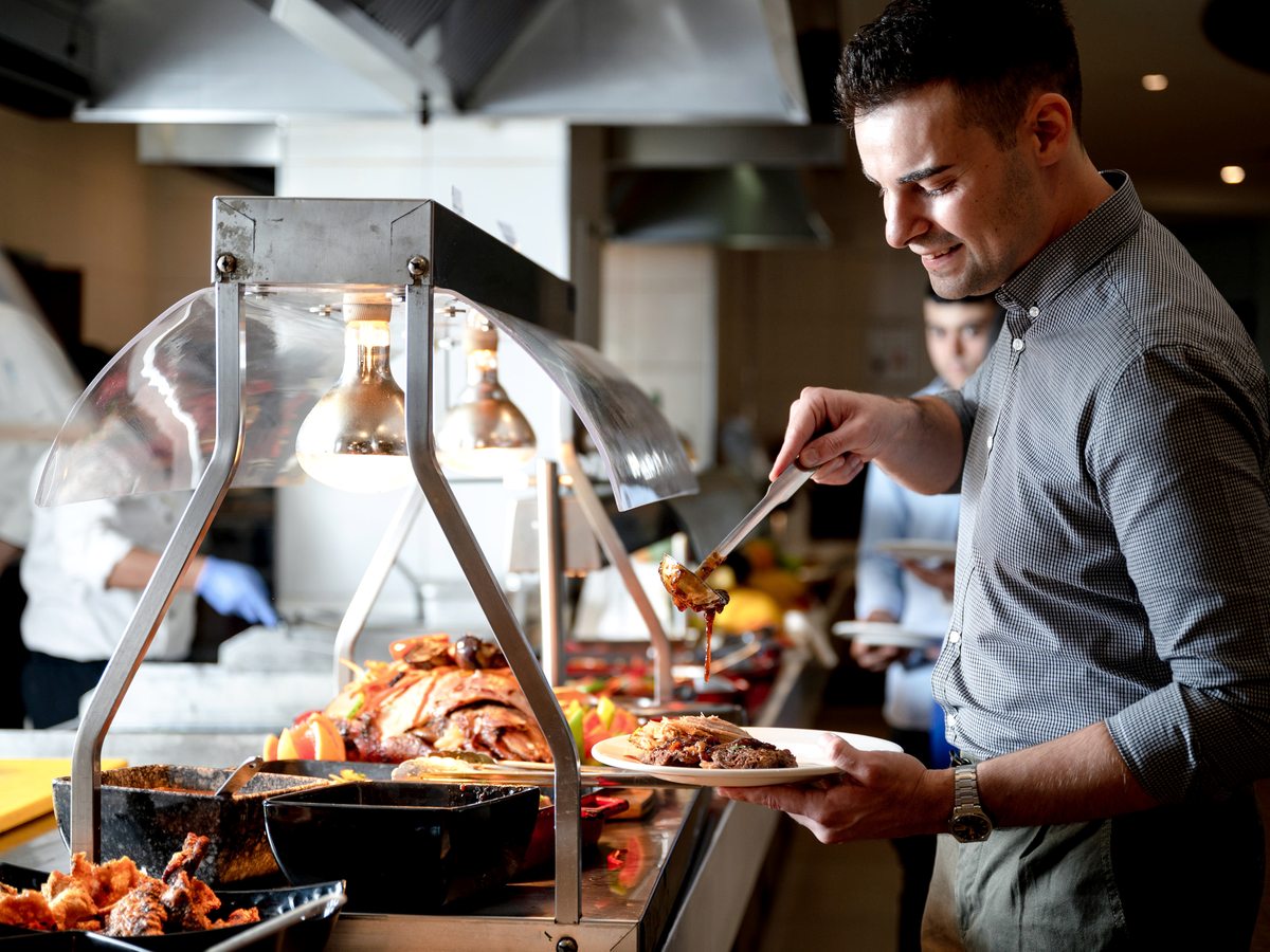 A guest serving from a hot buffet counter with a carving station and heat lamps in a self-service restaurant.