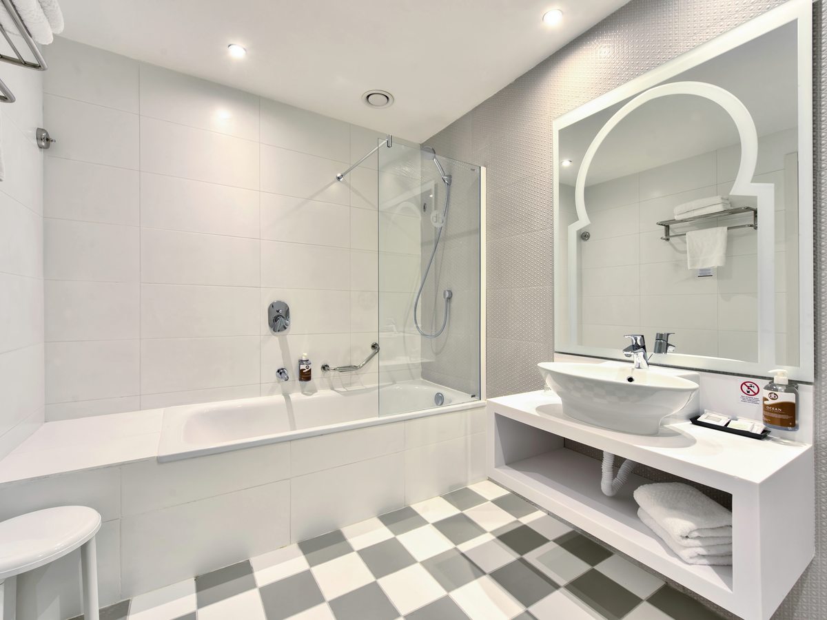A tiled bathroom with a bathtub under a glass shower screen, an open vanity washbasin, an arched mirror and a chequerboard floor.