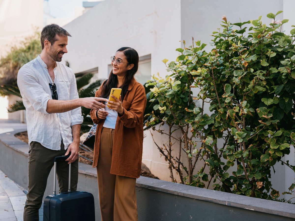 A couple with a wheeled suitcase on the hotel's entrance walkway, past raised planters and shrubs.