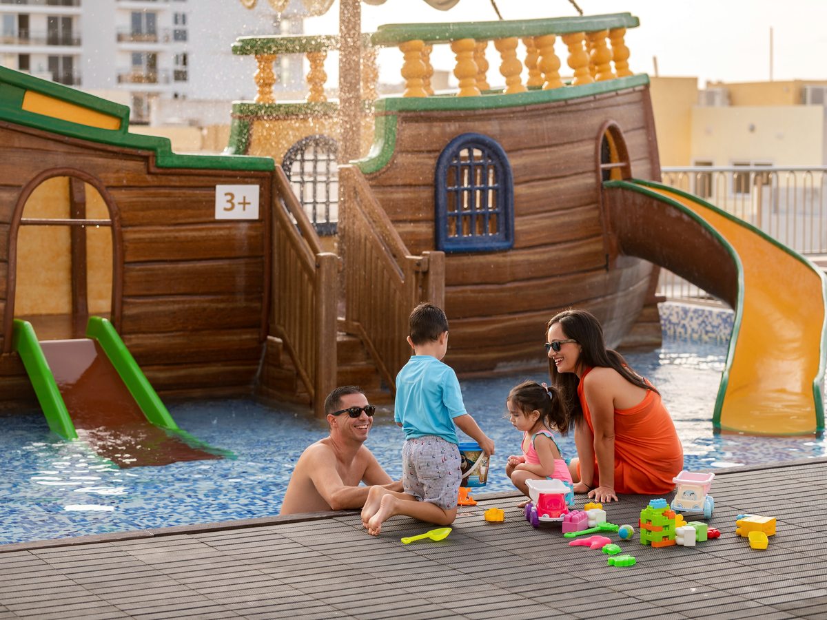 A large wooden pirate-ship play structure with green and yellow water slides, a family with two young children playing on the timber deck beside the shallow blue pool.