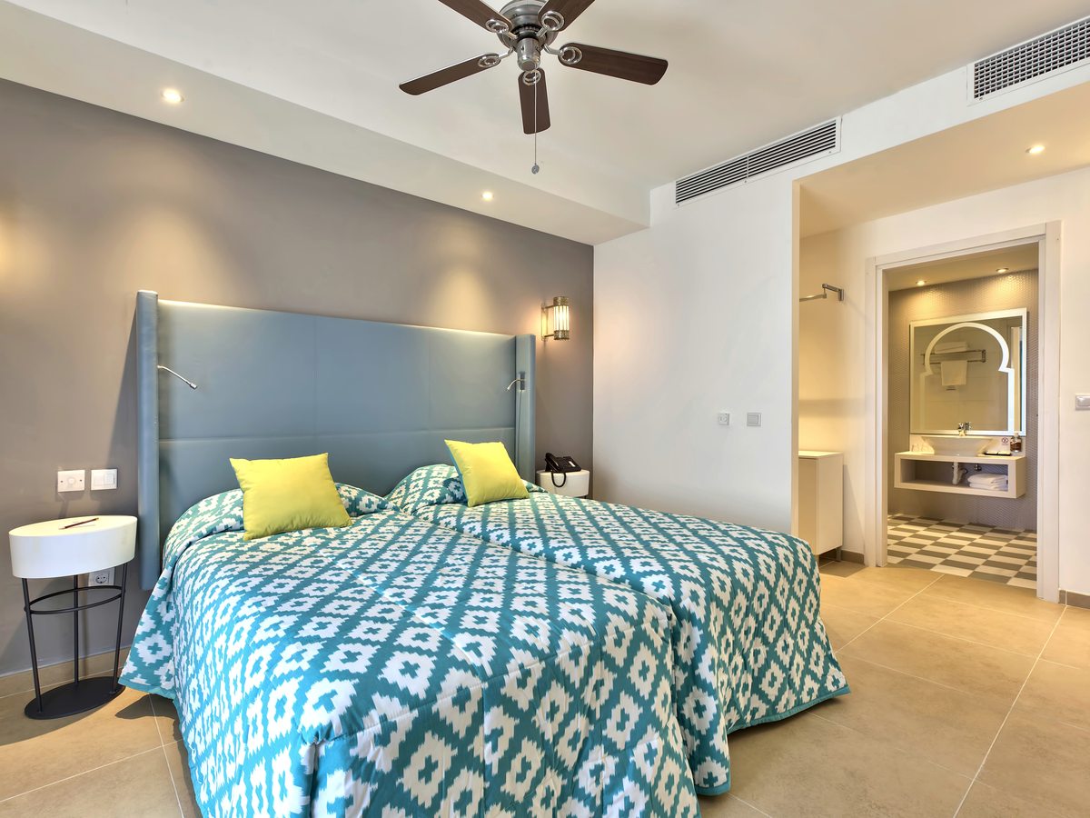 A room with two single beds and a padded headboard, an open doorway to the bathroom vanity, and a ceiling fan.