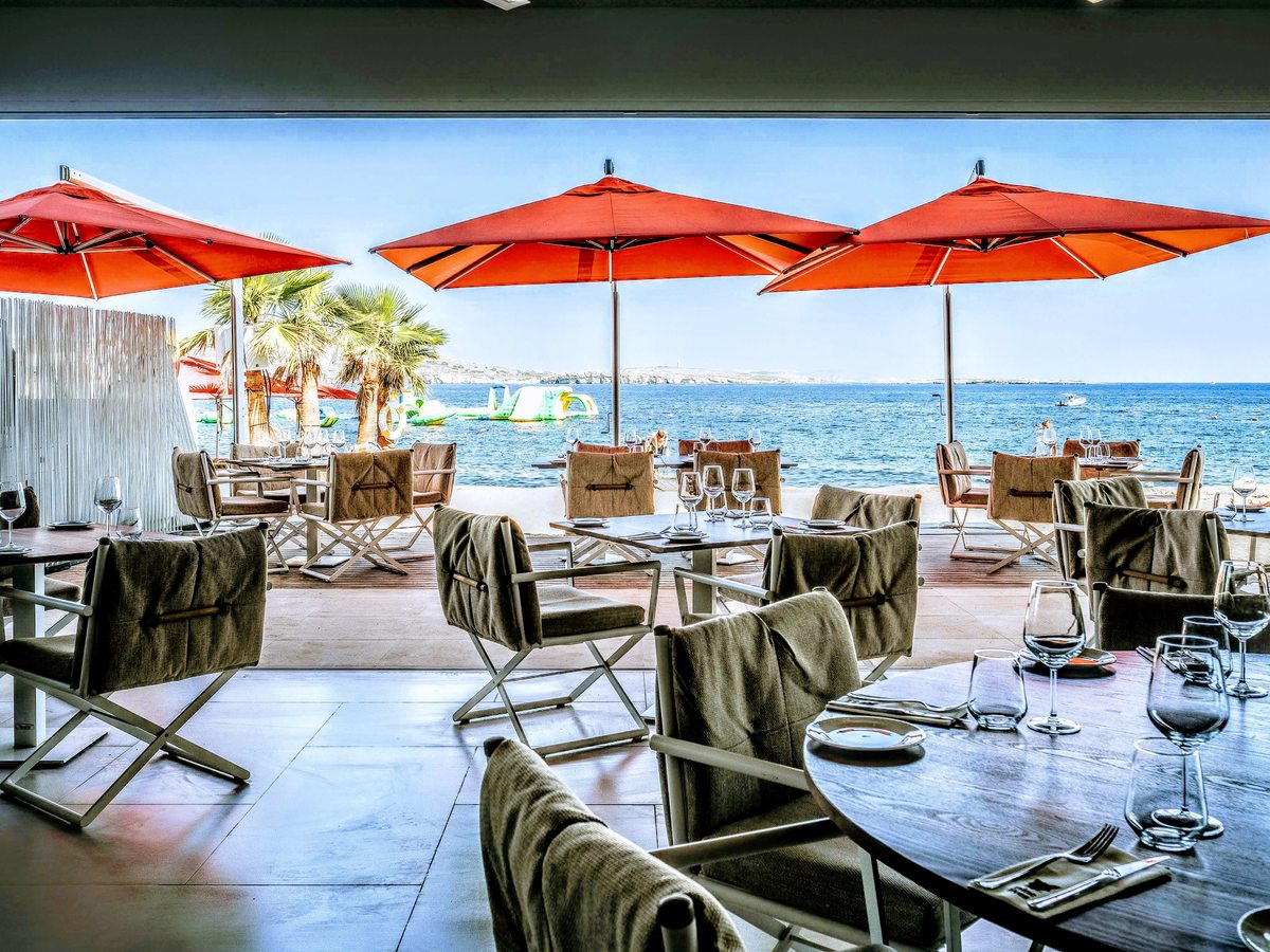 An open-air restaurant terrace with laid tables under red parasols and canvas shades, the sea beyond.