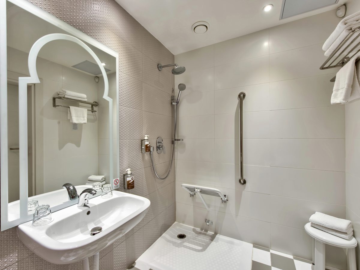 A step-free bathroom with a roll-in shower, a fold-down shower seat, grab rails, a wall-mounted basin and an arched mirror.