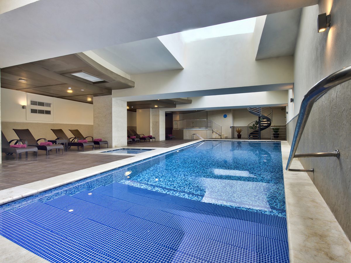 A rectangular indoor pool with blue mosaic tiles lit by a skylight, wicker loungers with pink towels along one side and a whirlpool alcove beyond.