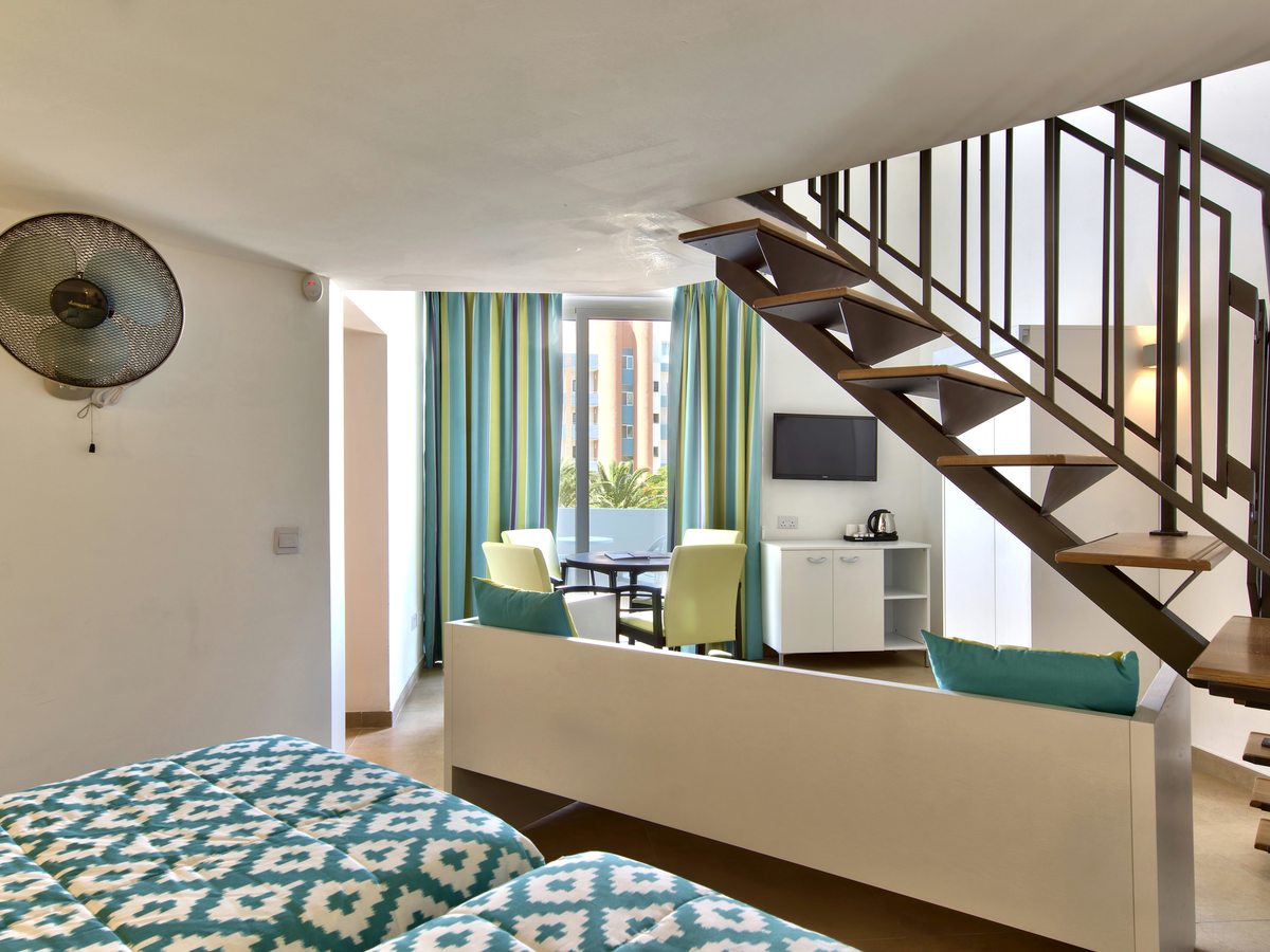 A split-level room with an internal metal-and-wood staircase to an upper level, a double bed below and a seating and dining area by the balcony.