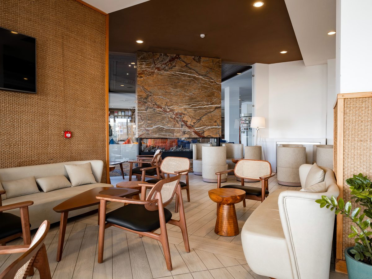 An indoor lobby lounge with cream sofas and timber-framed armchairs around low coffee tables, a marble feature wall and a wall-mounted television, on a tiled floor.